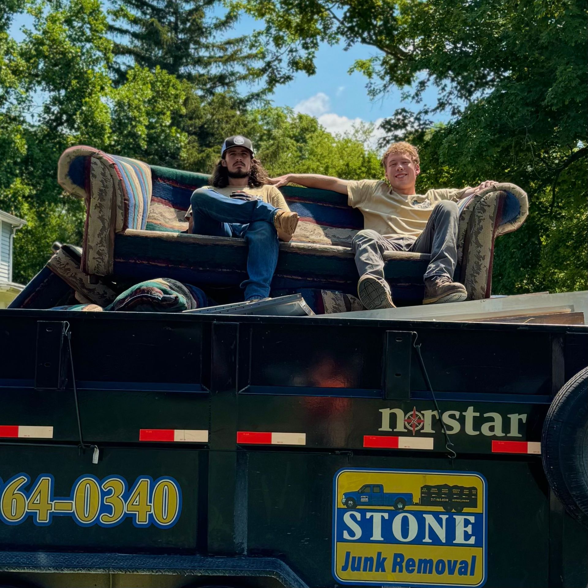 Two men sit on a damaged couch in a junk removal truck. Sunny day, trees in the background.