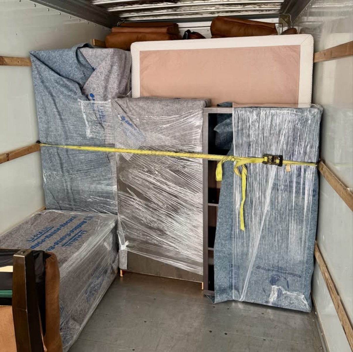 Two movers wrapping a chair in plastic wrap in a room with cardboard boxes and furniture.