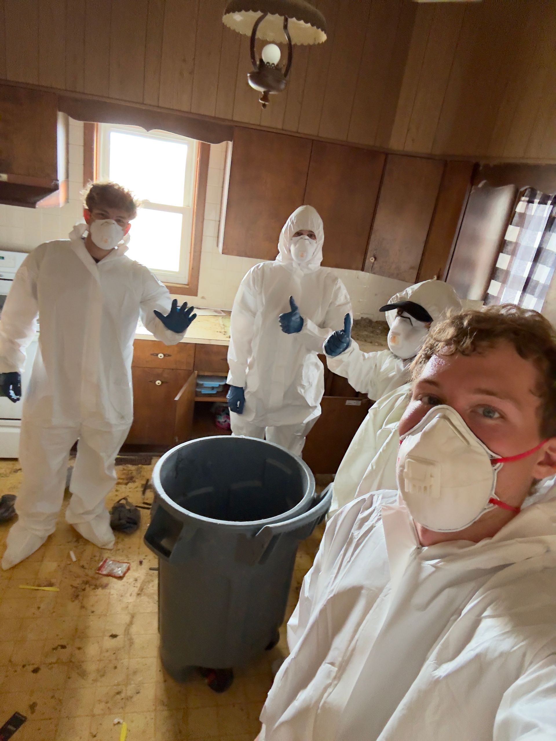 Four people in hazmat suits in a messy kitchen. One takes a selfie, others gesture. Gray trash can in foreground.