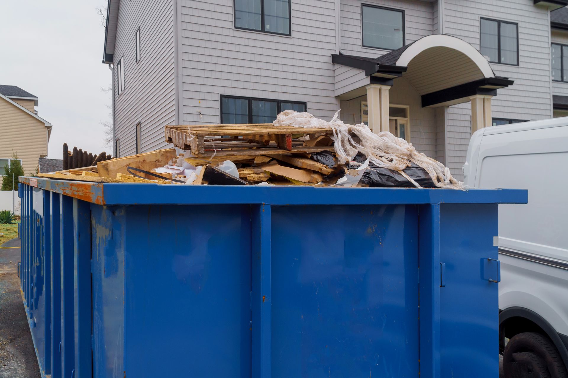 A large blue dumpster overflowing with construction debris and wood scraps outside a house.