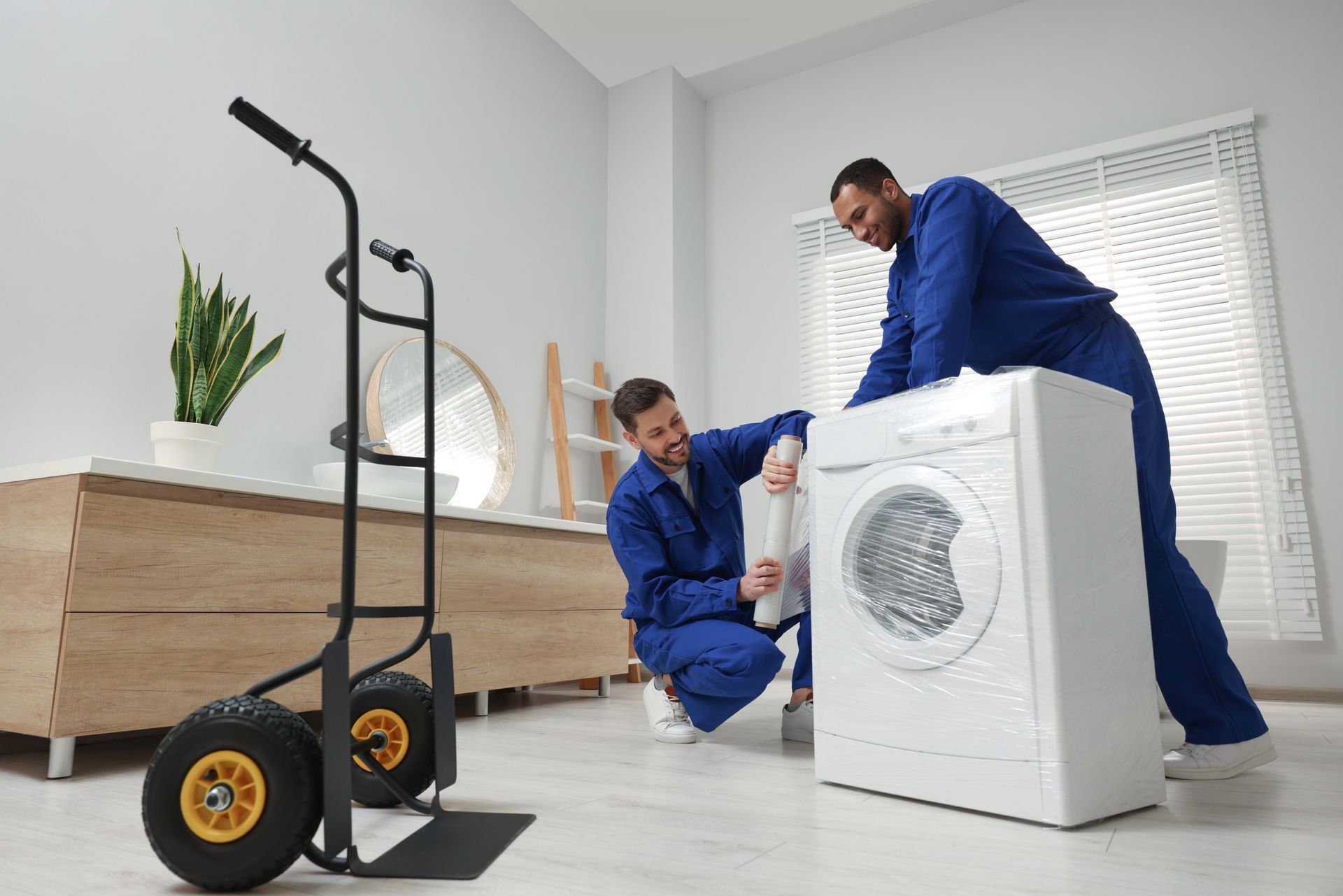 Two movers wrapping a washing machine indoors, near a hand truck.