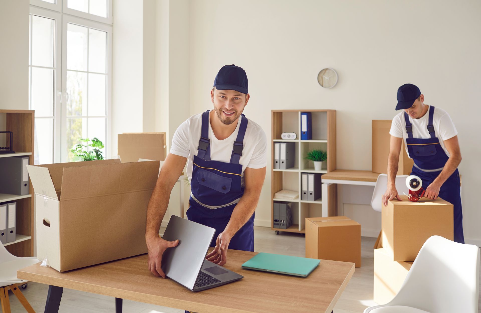 Two movers packing office belongings in a bright room with a laptop, boxes, and a desk.