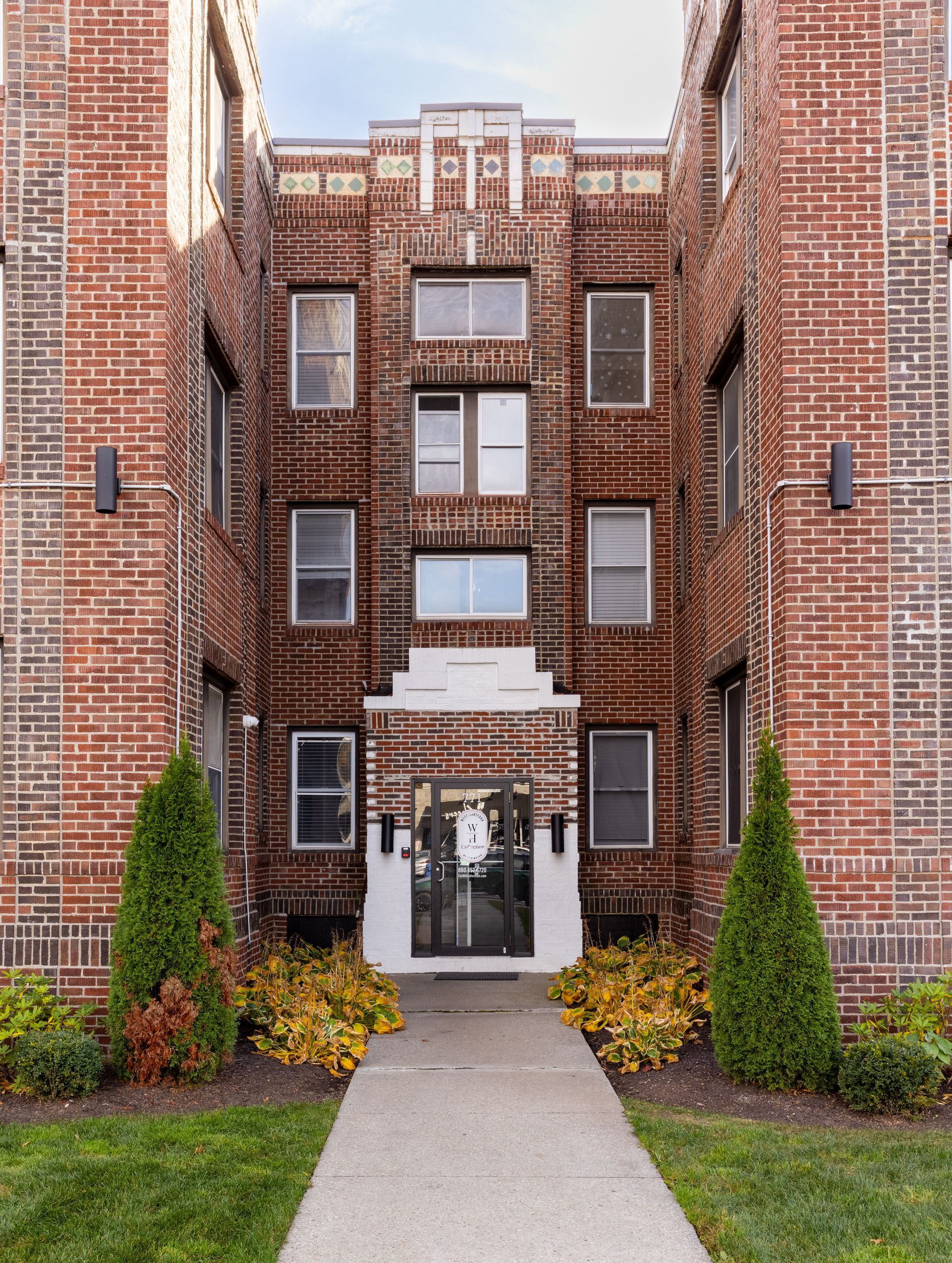 Brick apartment building entrance with a stone path, greenery, and a cross-shaped detail at the top.