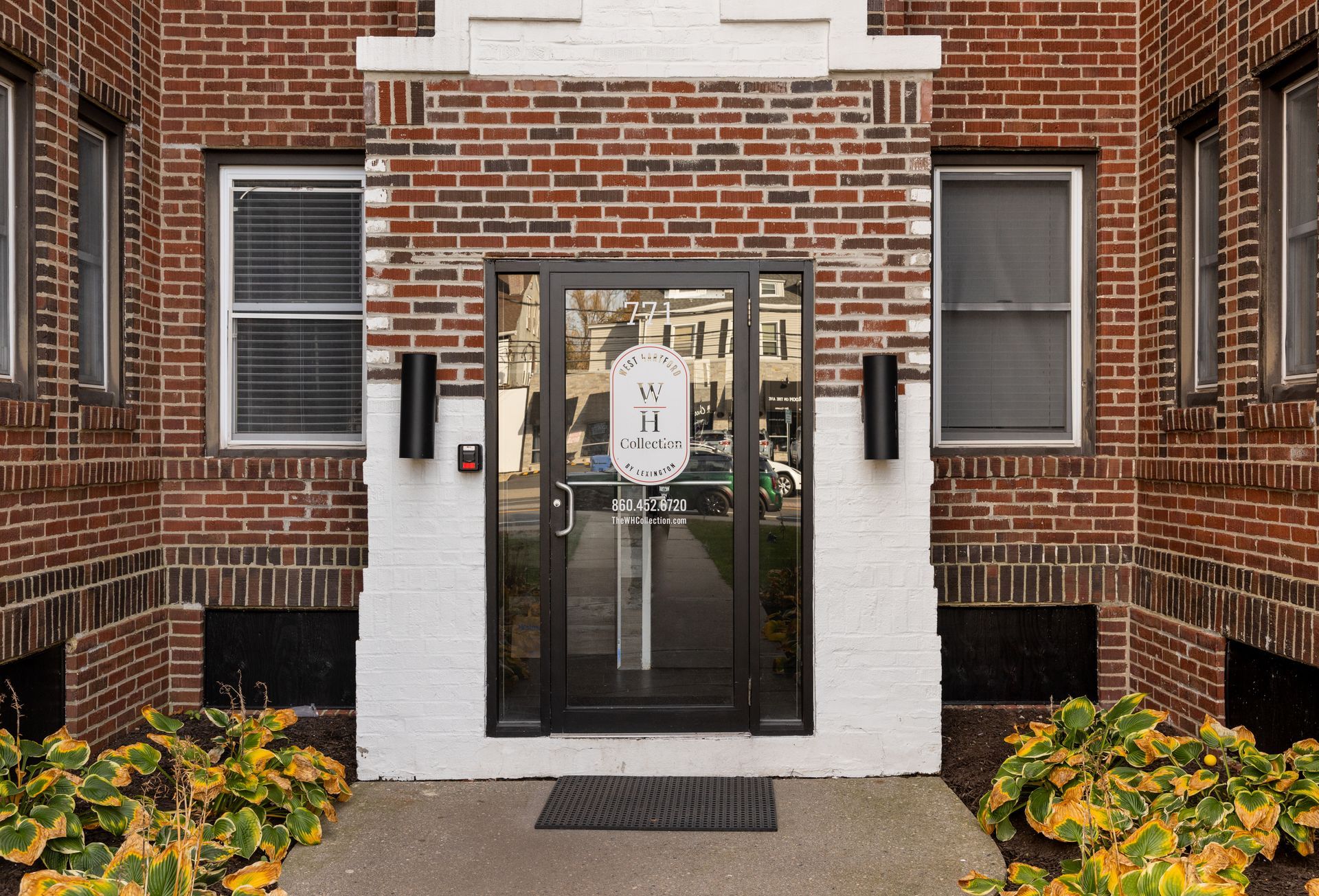 Brick building entrance with glass door and address, flanked by windows and bushes.