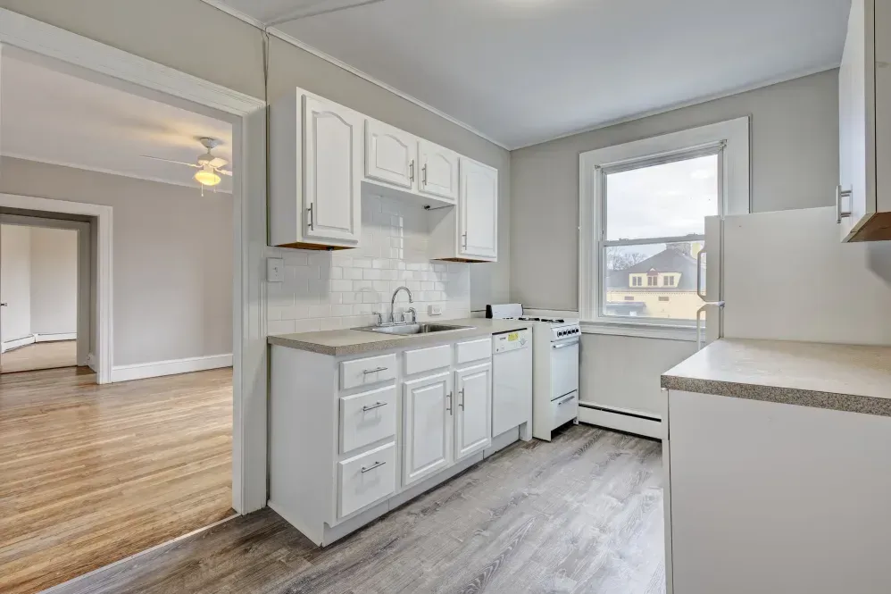 Bright kitchen with white cabinets, countertop, and appliances. Gray walls, window, and flooring. Opens to a living area.