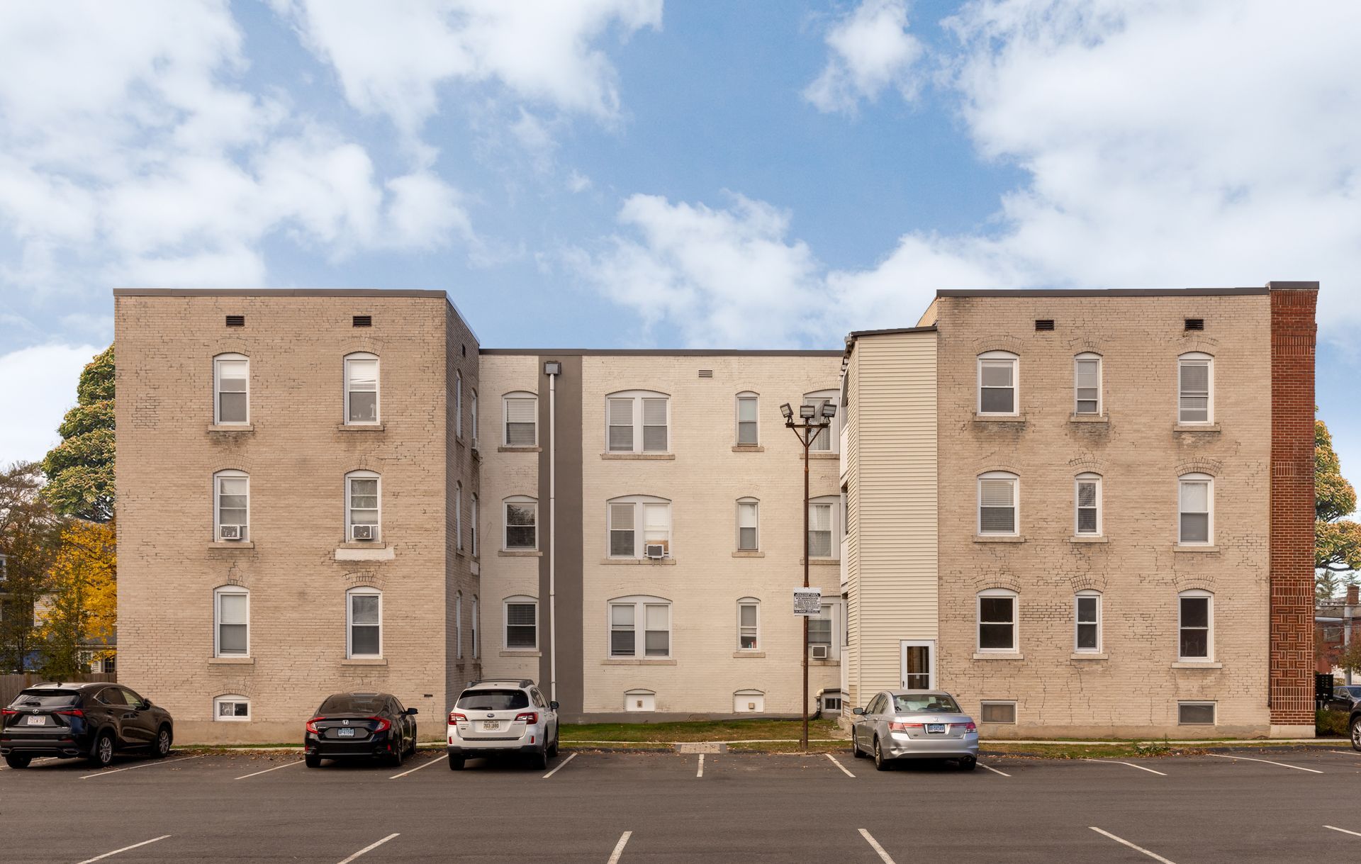 Multi-story apartment building with parked cars in front. Light brick and beige exterior. Blue sky.