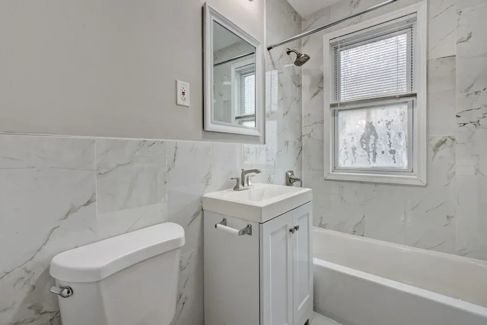 White bathroom with a toilet, sink, mirror, and tub, with marble-look tiles.
