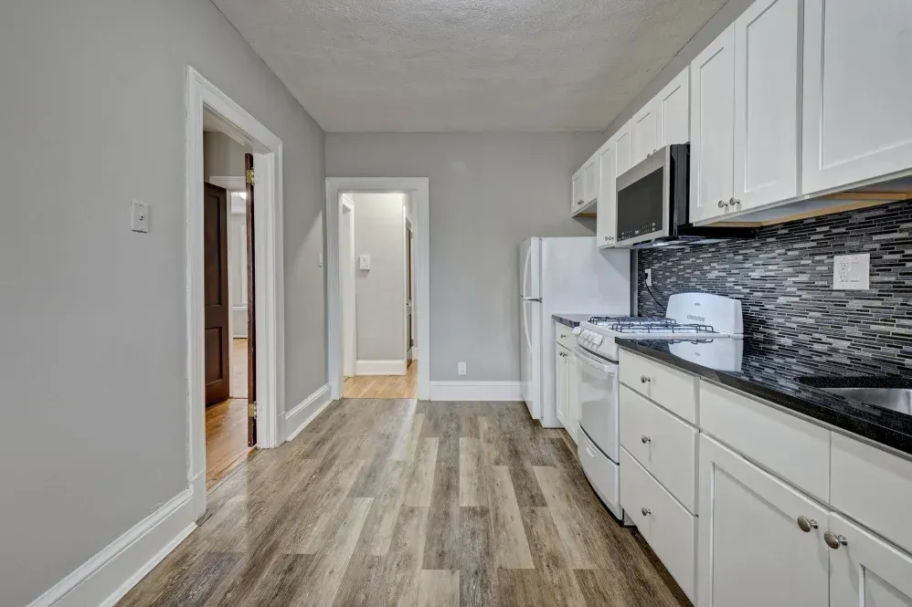 Narrow kitchen with white cabinets, appliances, and gray walls; wood-look floor.