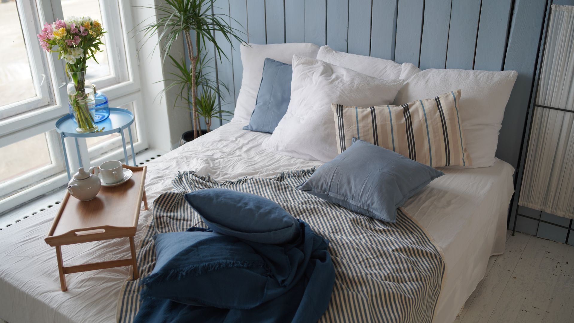Cozy bedroom with blue accents: bed with pillows, tray with tea, flowers by window.