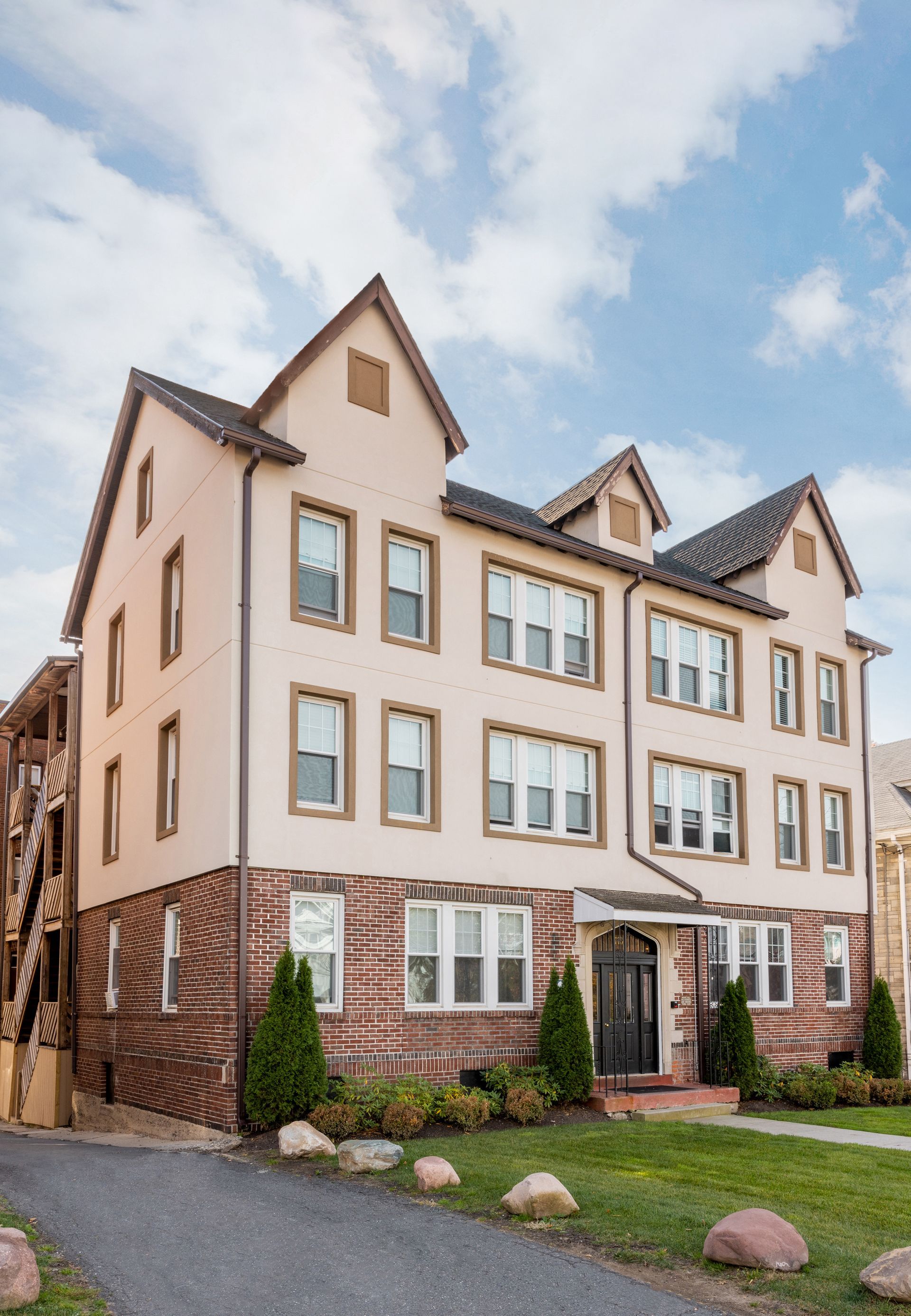 Multi-story apartment building with brick base, beige stucco, and gabled roof under a cloudy sky.