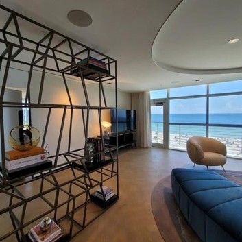 A modern living room with a geometric metal shelf, a blue ottoman, an armchair, and a large window overlooking the ocean.
