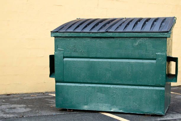 A green dumpster is sitting in a parking lot in front of a yellow wall.