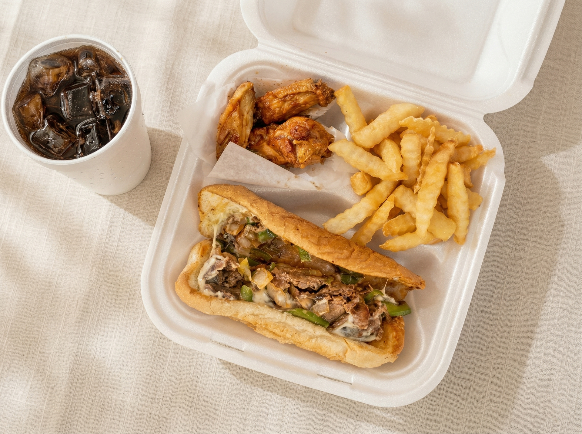 A takeout container holds a Philly cheesesteak, crinkle-cut fries, and fried chicken, with a cup of dark soda to the side.