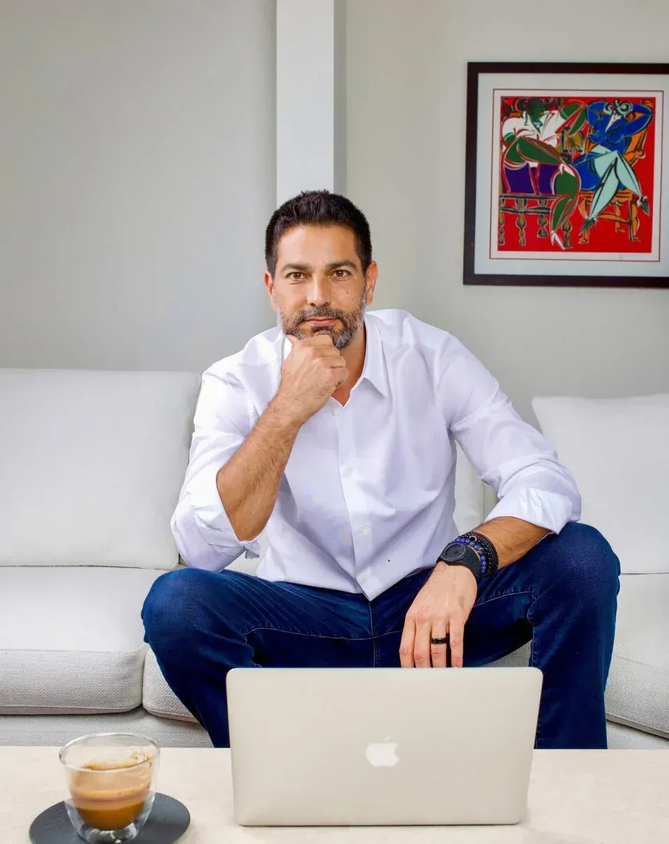 Man in white shirt and jeans sits on a sofa, looking at a laptop. Hand on chin. Coffee cup on table. Painting on wall.