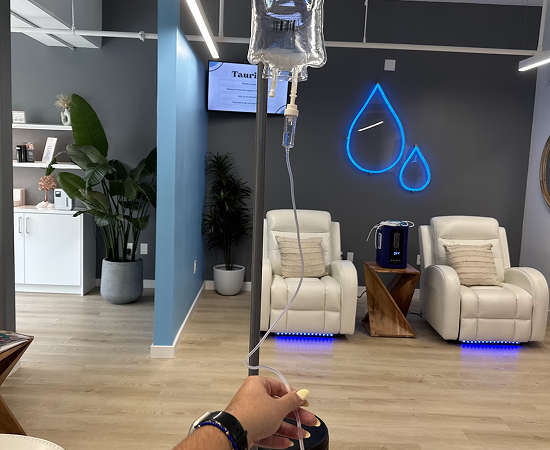 IV drip in progress, modern spa setting. Reclining chairs, neon water drop sign, and interior plants visible.