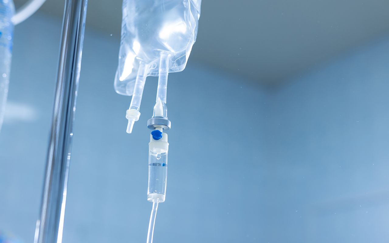An IV drip bag and drip chamber hanging from a medical stand against a blurred blue background.