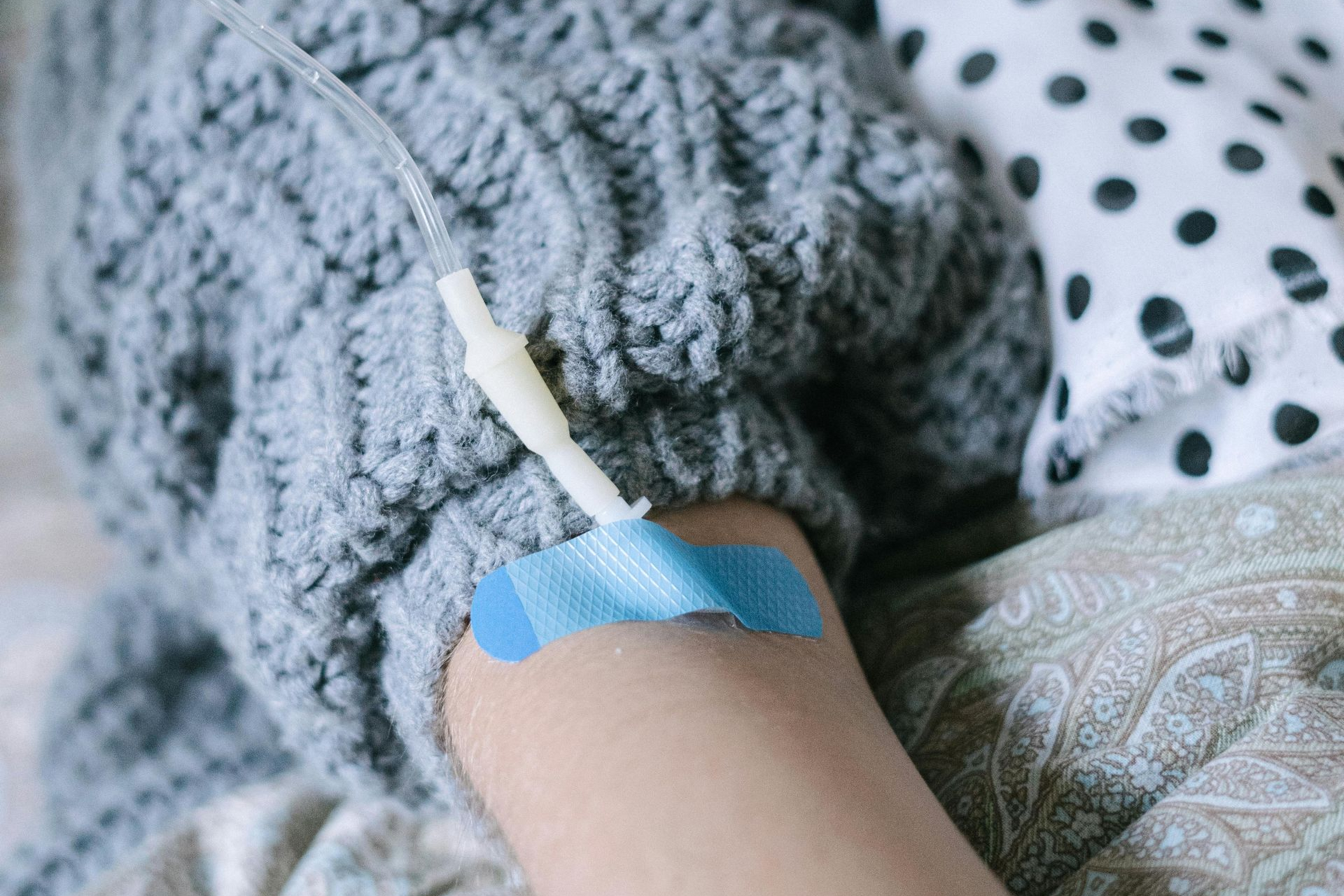 A person's arm with an IV line secured by blue medical tape, resting against a textured gray sweater and patterned pillow.