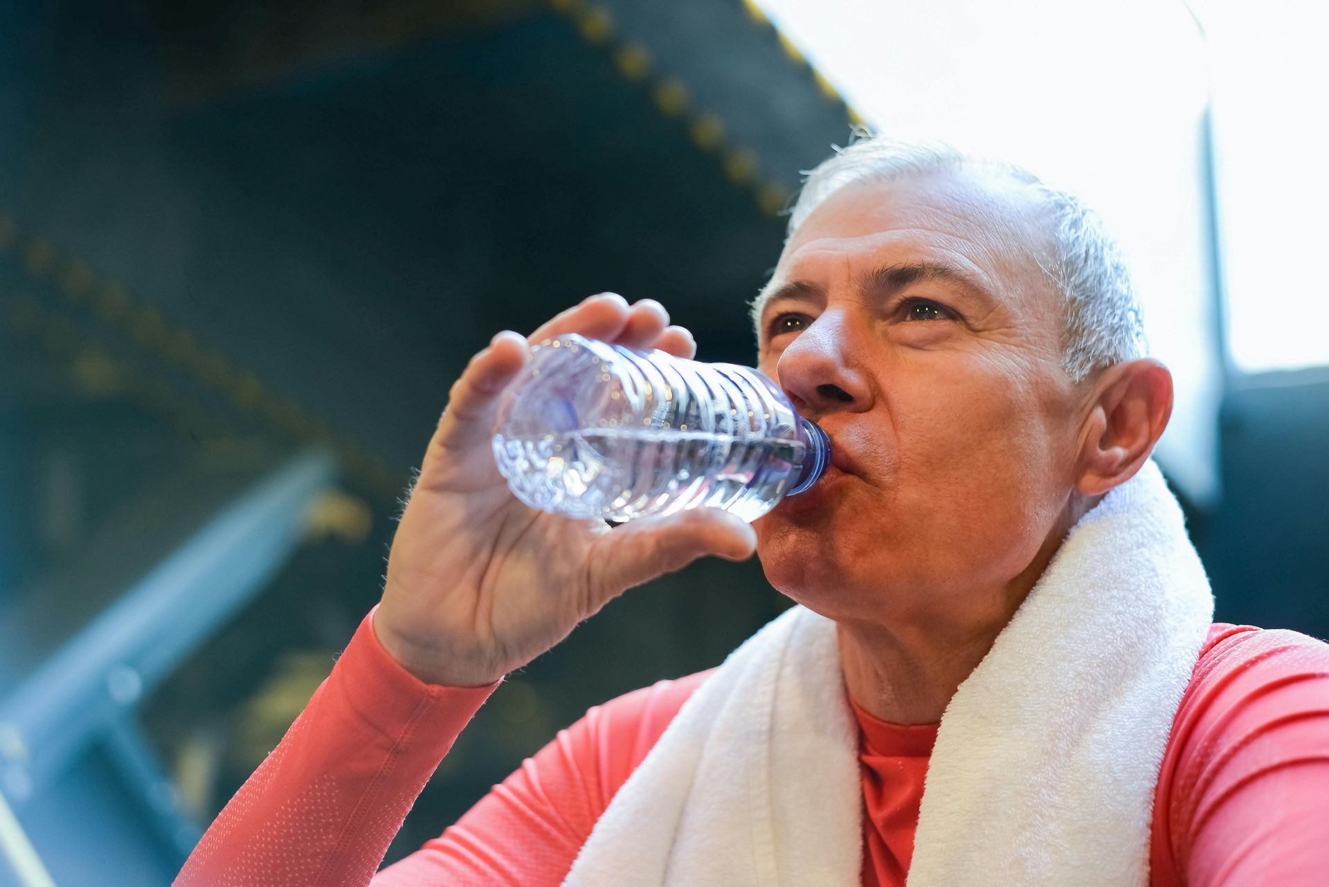 A person in a coral-colored athletic shirt with a white towel around their neck drinks from a plastic water bottle.