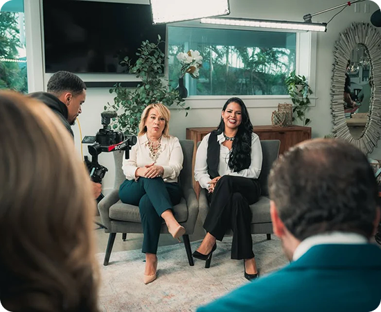 Two women sit for an interview with camera operator. Interior, bright light, neutral-toned room.
