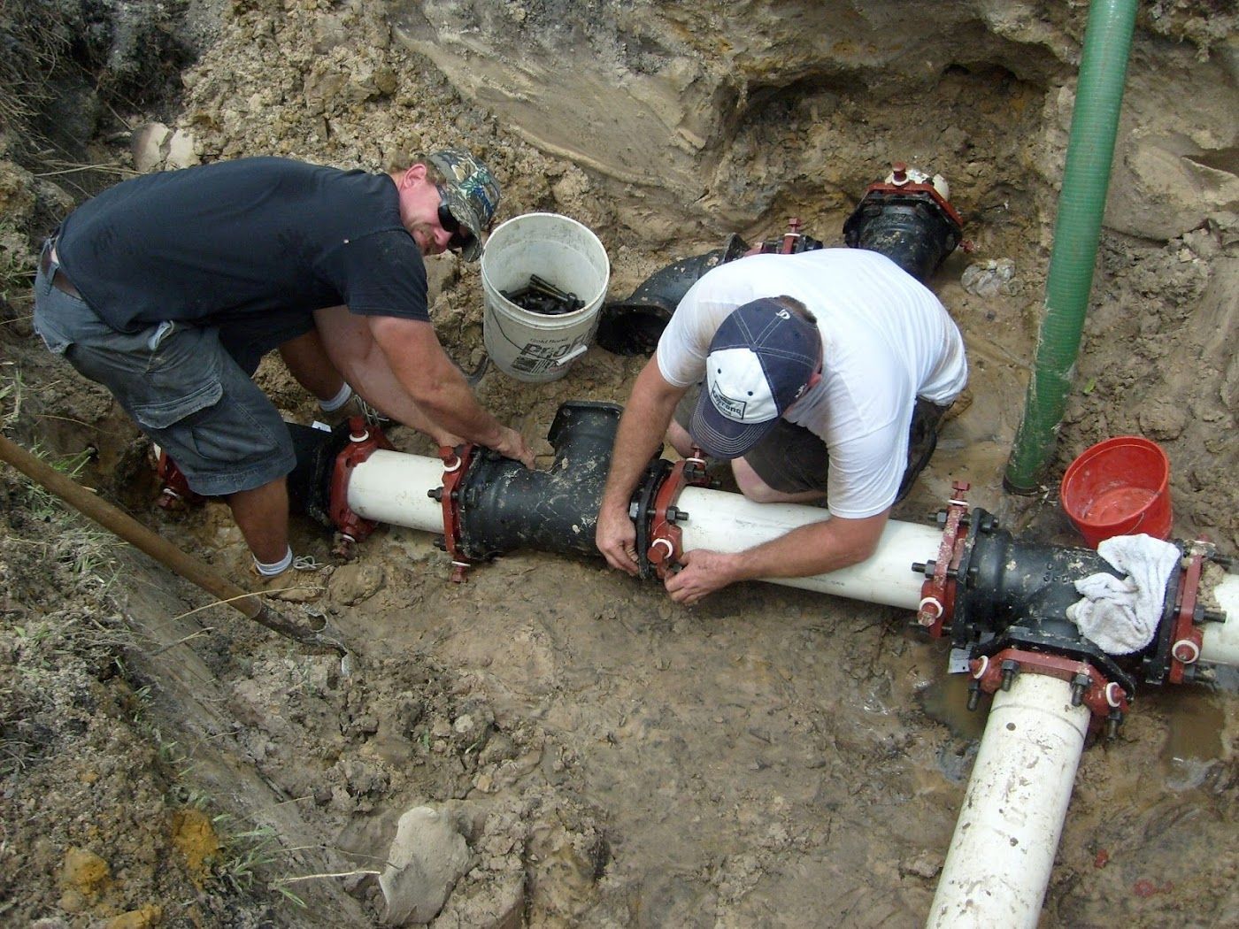 Two workers installing water pipes in a dirt trench, using tools and fasteners.