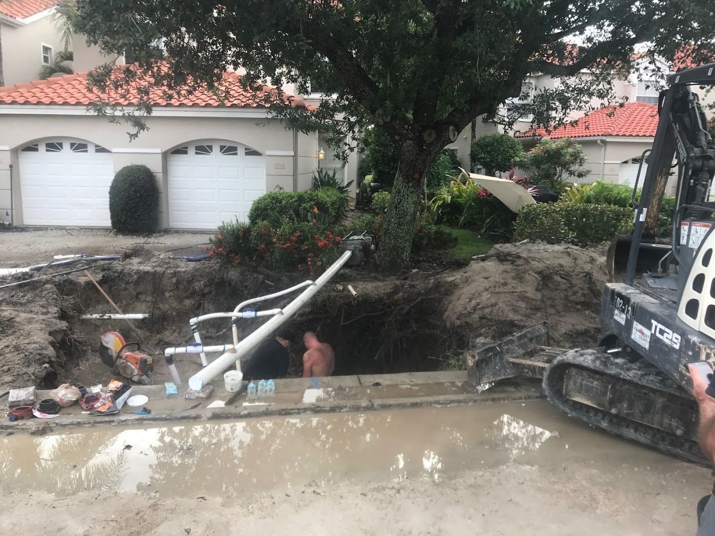 Excavator next to a dug-up swimming pool. Person in hole with pool equipment and debris. Houses in background.
