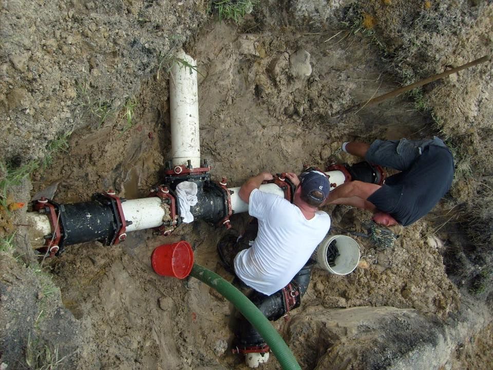 Two people repairing underground pipes in a dirt trench, one in a white shirt, using tools.
