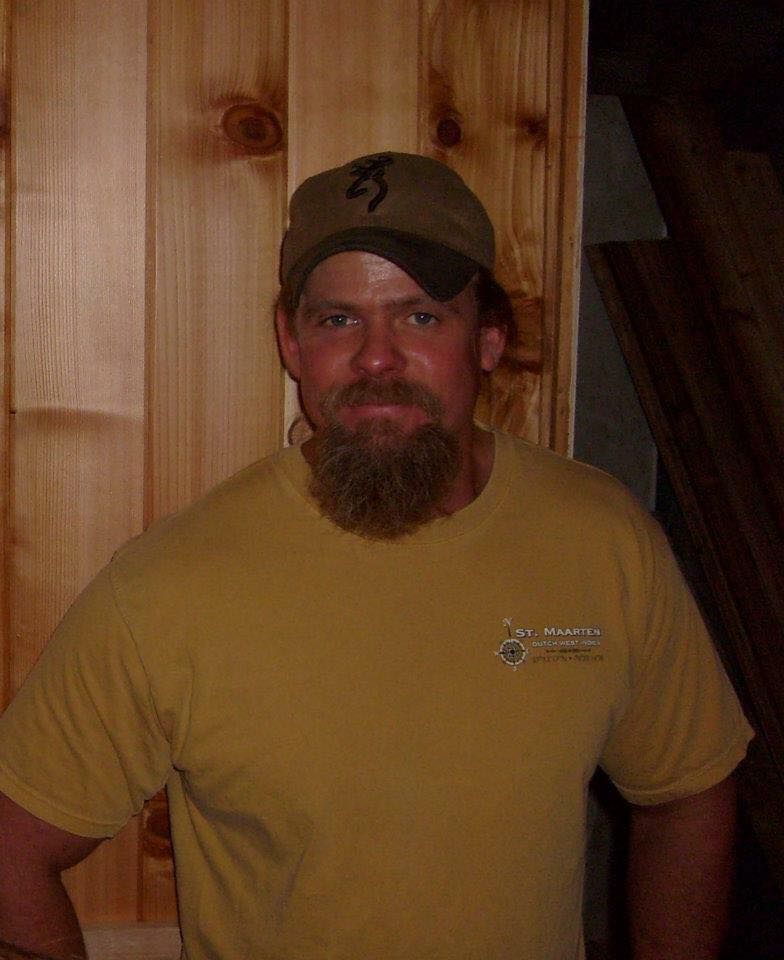Man with beard wearing a baseball cap and yellow shirt, standing against wood paneling.