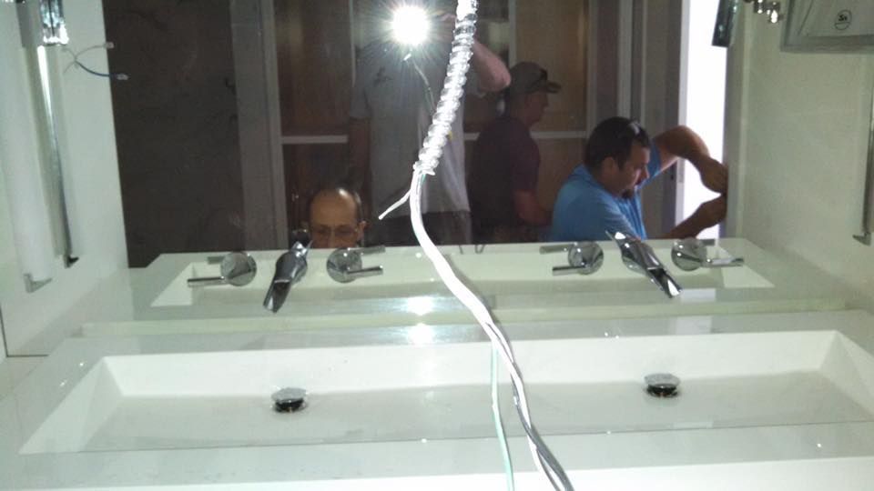Two sinks with workers in the background; a bathroom under construction with bright lights and mirrored walls.