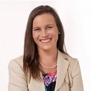 A smiling professional wearing a tan blazer over a multi-colored floral top and a pearl necklace against a white background.