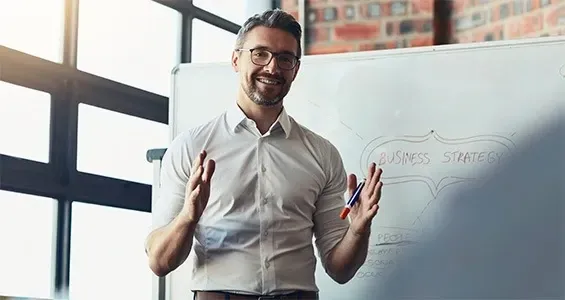 A person in a white button-down shirt stands before a whiteboard, gesturing while presenting in an office setting. A person in a white button-down shirt stands before a whiteboard, gesturing while presenting in an office setting.