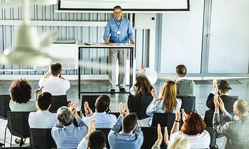 A speaker stands at a podium in front of a conference room filled with seated attendees listening to a presentation.