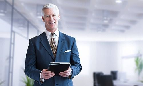 A professional in a suit holds a tablet while standing in a bright, modern office.
