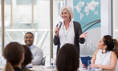 A professional speaker holding a microphone addresses a small, attentive group in a brightly lit office meeting room.
