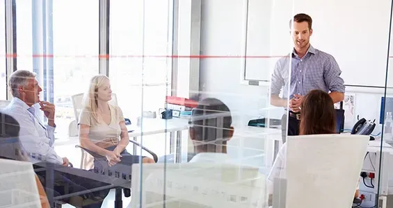 A man standing and presenting to a group of colleagues sitting around a table in a modern, glass-walled office. A man standing and presenting to a group of colleagues sitting around a table in a modern, glass-walled office.