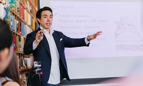A speaker gesturing in front of a presentation screen in a room with bookshelf walls.
