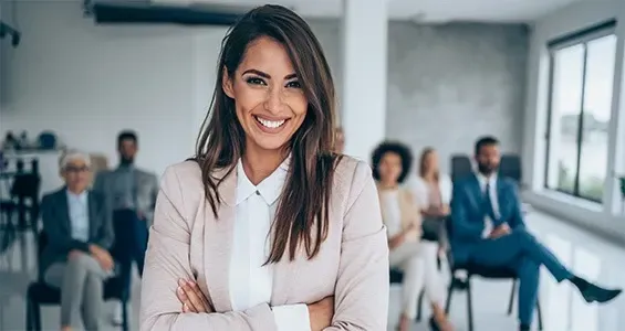 A person with long dark hair and a beige blazer stands smiling in the foreground, with colleagues sitting in an office. A person with long dark hair and a beige blazer stands smiling in the foreground, with colleagues sitting in an office.