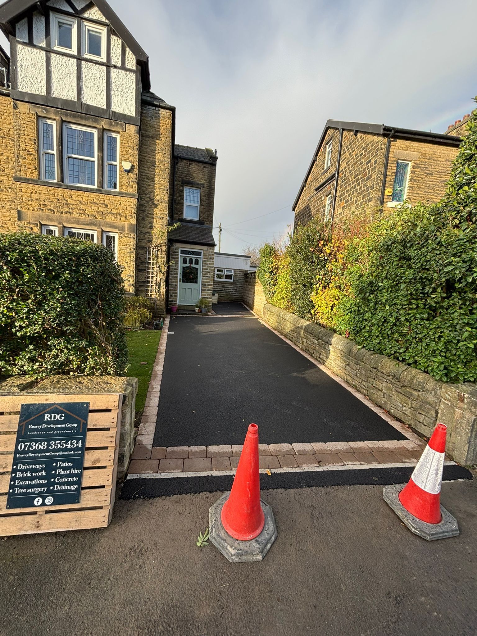 Driveway entrance between stone buildings, blocked by two orange cones and a low barrier.