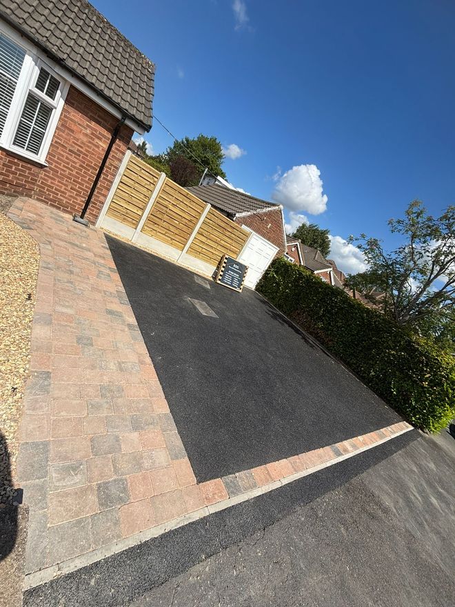 Tilted view of a suburban driveway with brick houses, a black carport, a hedge, and blue sky