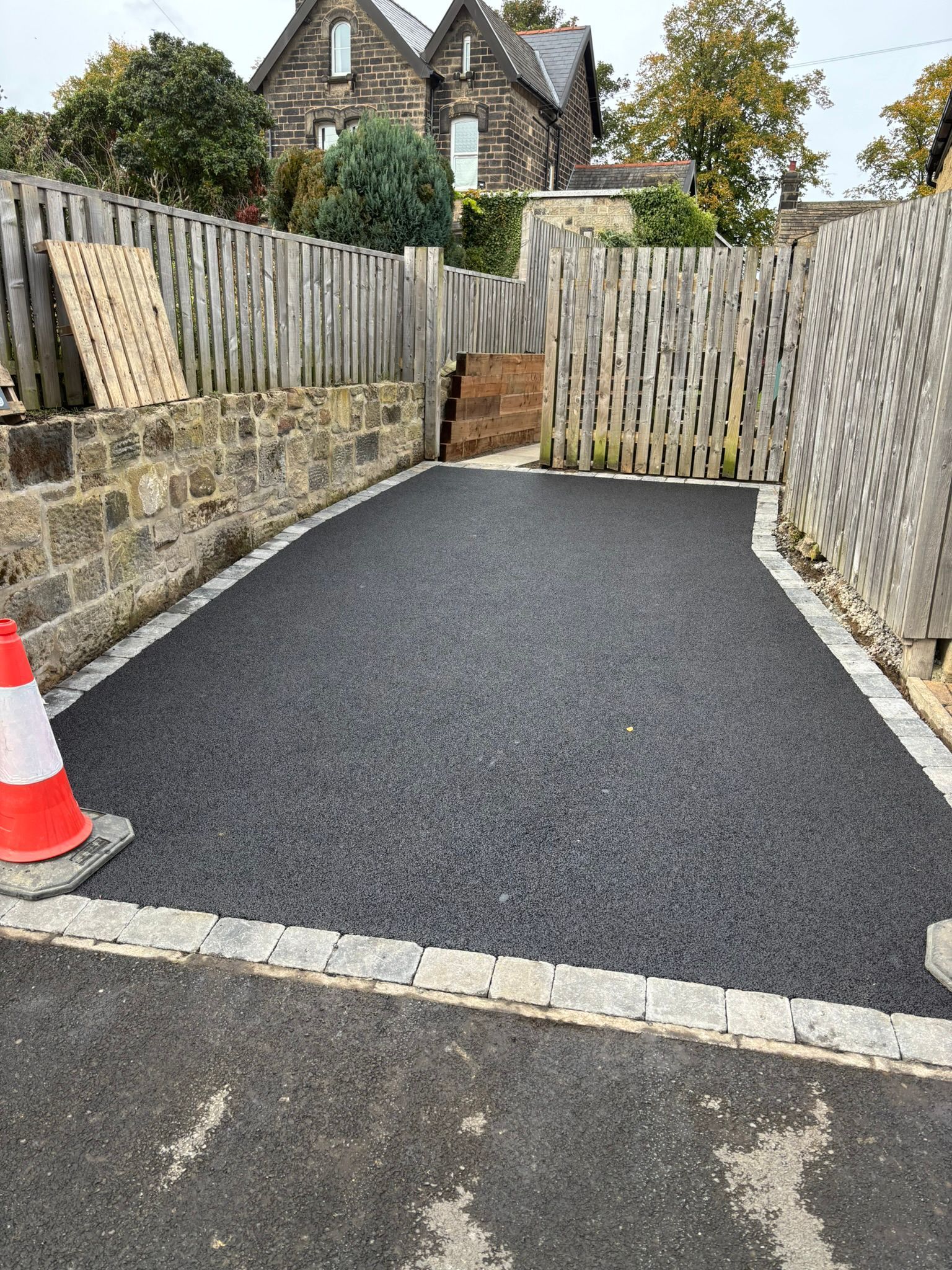 Freshly paved driveway with a cone, stone walls, wooden fences, and a house in the background