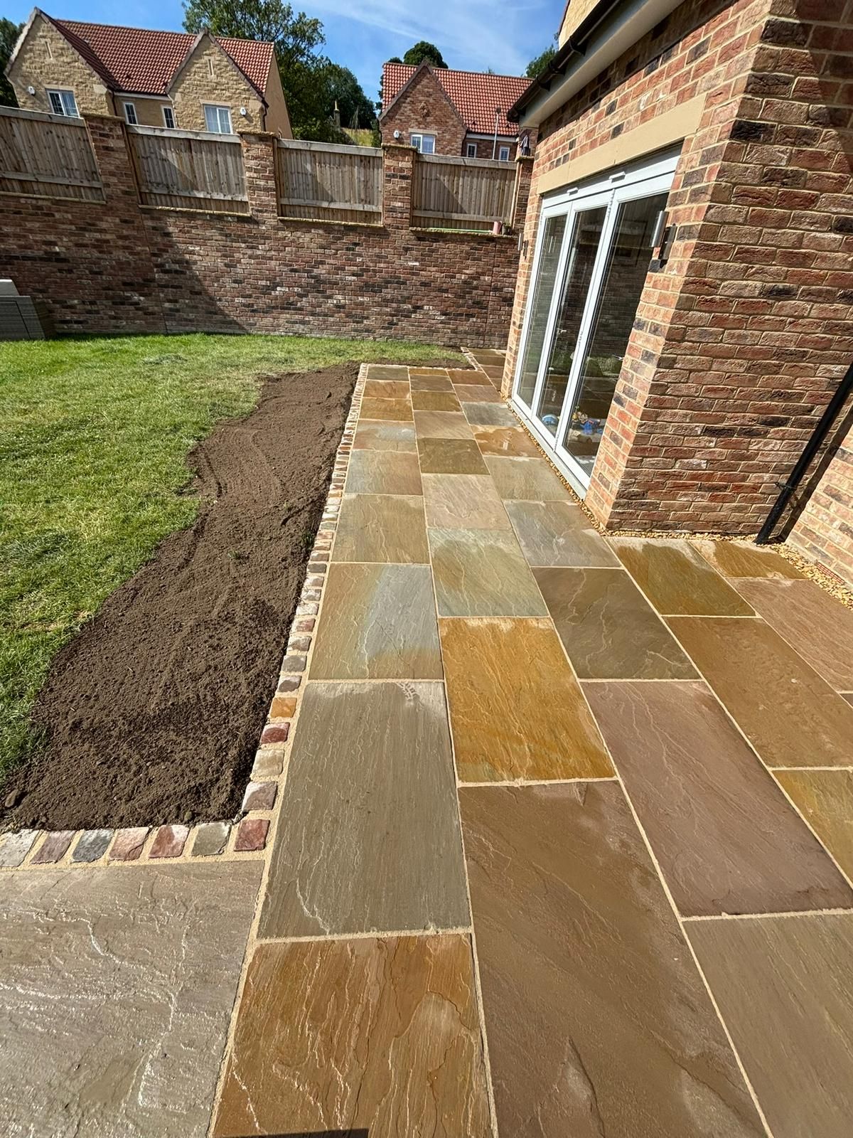 Stone patio beside a lawn and brick house, leading to sliding glass doors in a backyard.