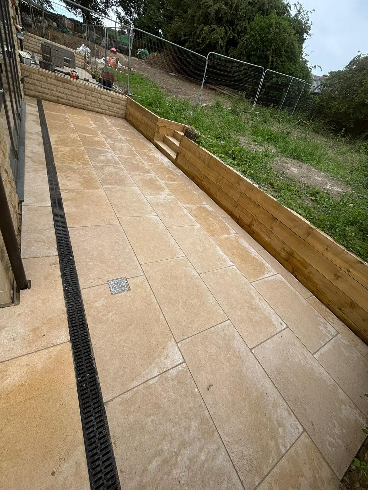 Paved outdoor ramp with tan tiles, black drainage strip, and raised retaining walls beside a grassy yard