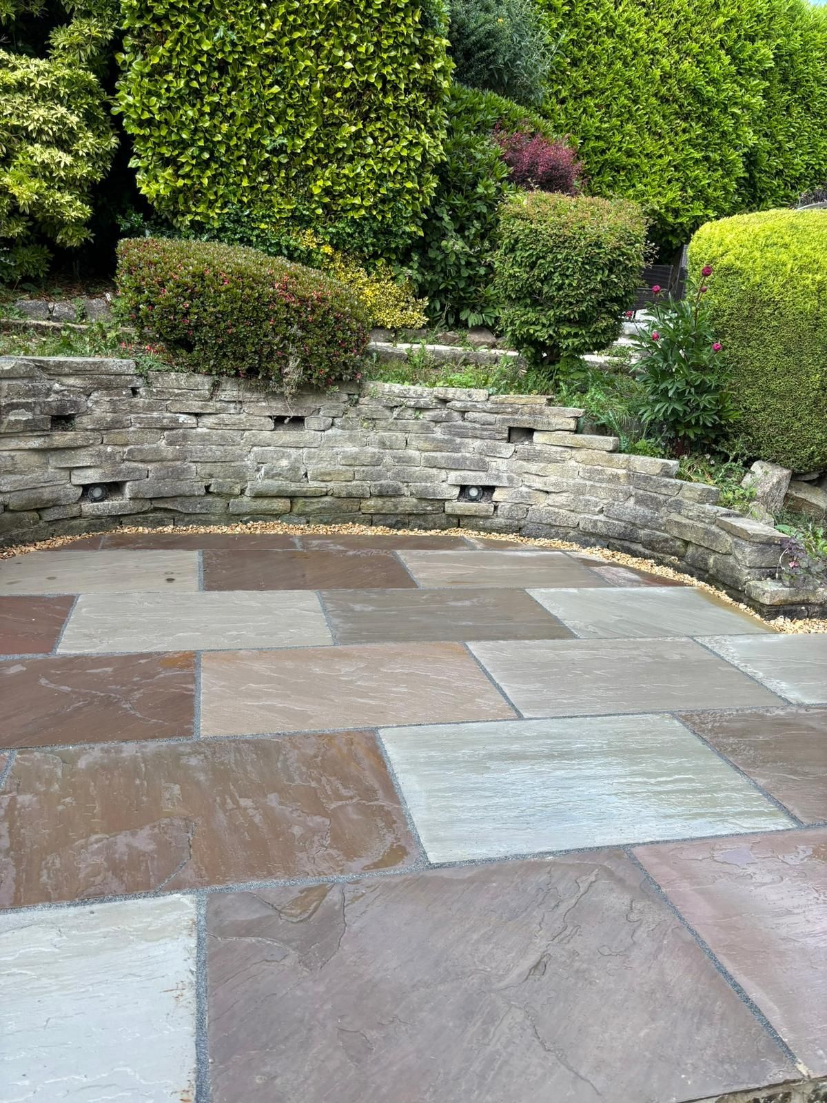Stone patio with multicolored tiles, low brick wall, and manicured green shrubs in a garden