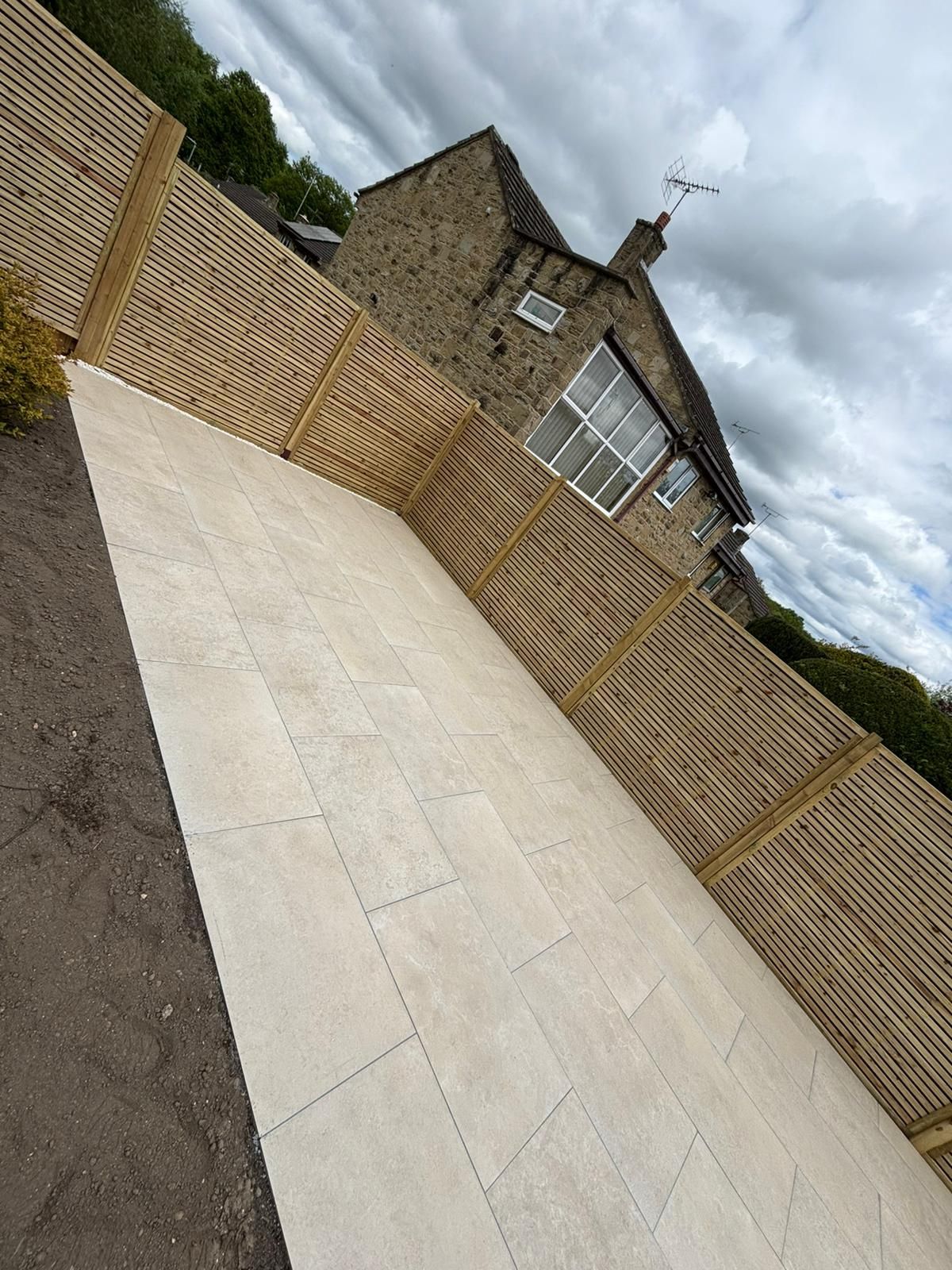Tilted view of a paved driveway, brick house, and wooden fence under a cloudy sky.