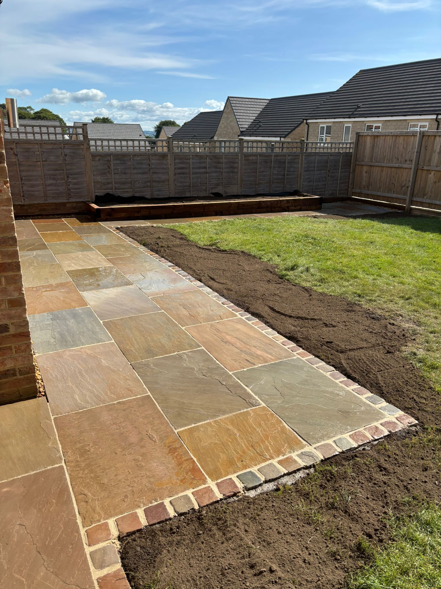 Backyard patio with paved stone walkway, grass, and wooden fence under a partly cloudy sky