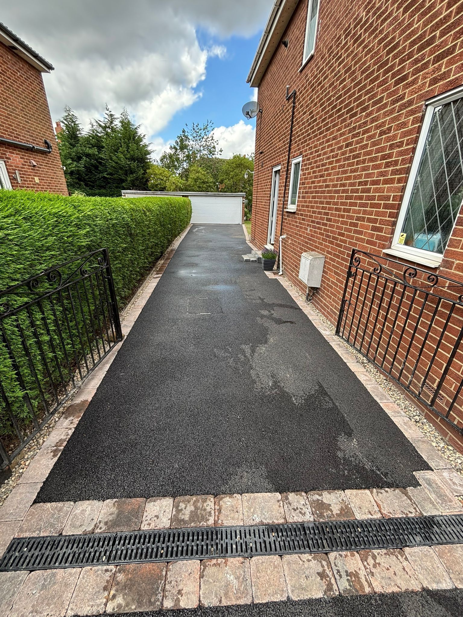 Narrow paved path between brick houses, with hedge on the left and a drain grate across the foreground.