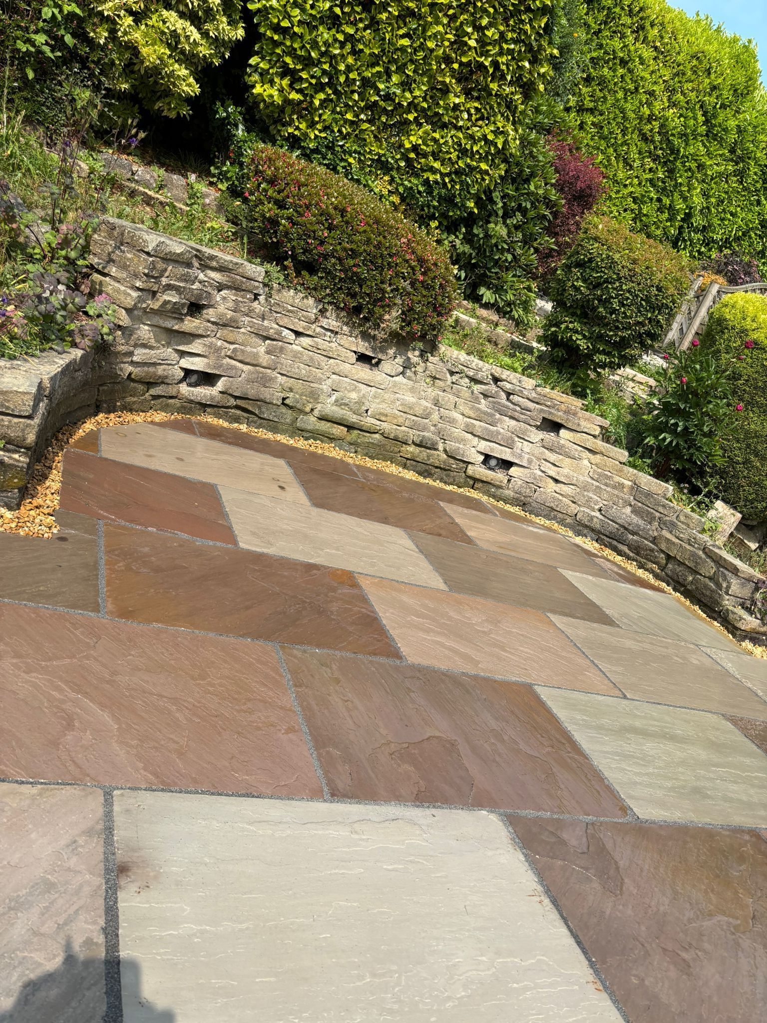 Stone patio beside a low rock wall and green shrubs, with a patchwork of tan and brown paving stones.