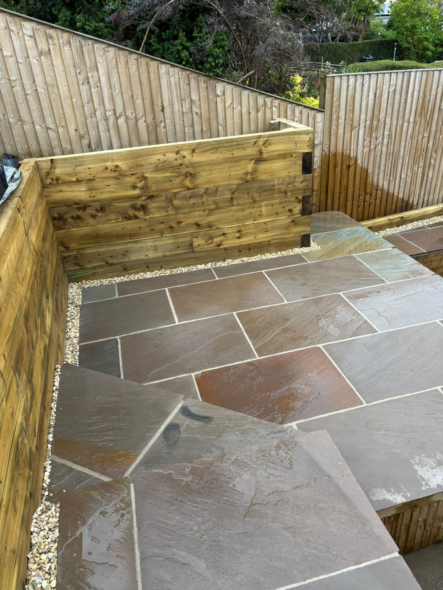 Outdoor patio with stone paving, timber retaining walls, and a fenced garden backdrop