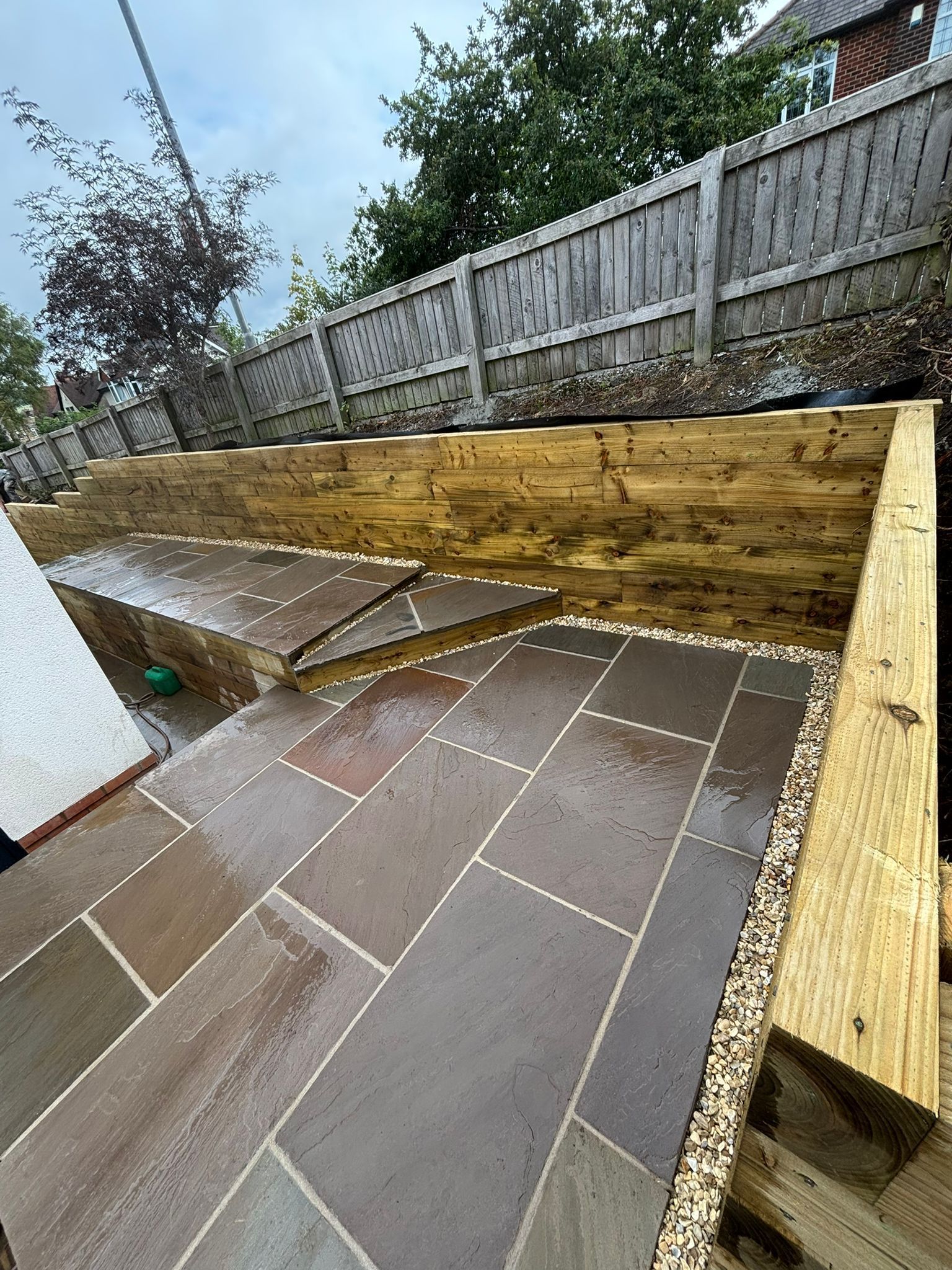 Wet tiled patio under construction with wooden retaining wall and fence along the edge