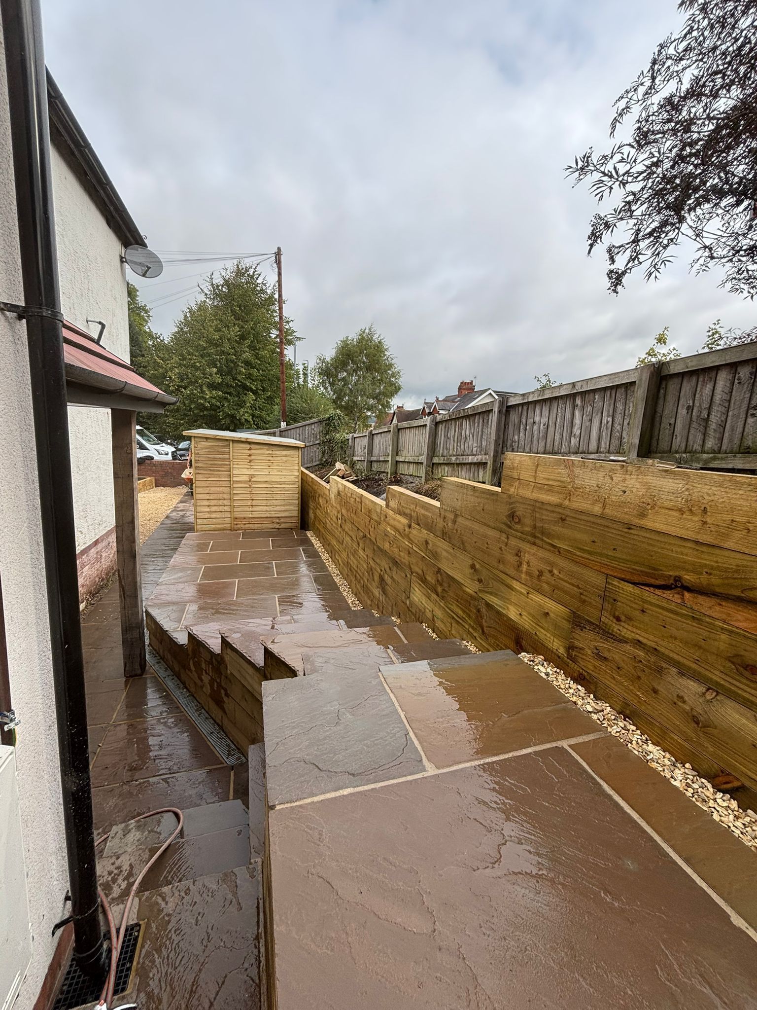 Waterlogged backyard walkway beside a house, with stacked pallets and floodwater pooling along a fence.