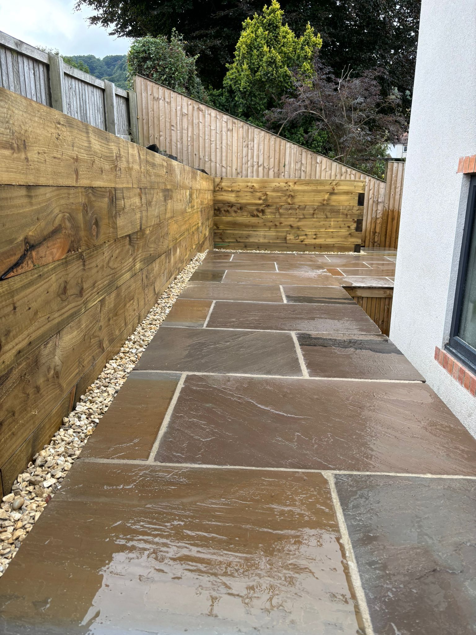 Wet stone patio walkway beside a house, with a fenced retaining wall and steps at the end.